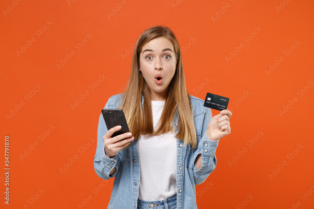 Shocked amazed young woman girl in casual denim clothes posing isolated on orange wall background studio portrait. People lifestyle concept. Mock up copy space. Using mobile phone, credit bank card. Shocked amazed young woman girl in casual denim clothes posing isolated on orange wall background studio portrait. People lifestyle concept. Mock up copy space. Using mobile phone, credit bank card.