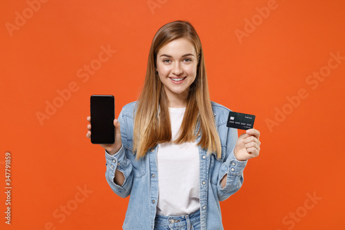 Smiling young woman girl in casual denim clothes posing isolated on orange wall background. People lifestyle concept. Mock up copy space. Hold mobile phone with blank empty screen, credit bank card.