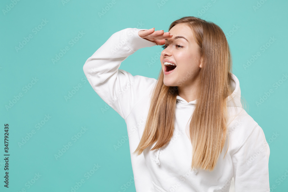 Excited young woman girl in casual white hoodie posing isolated on blue turquoise background studio. People lifestyle concept. Mock up copy space. Holding hand at forehead looking far away distance. Excited young woman girl in casual white hoodie posing isolated on blue turquoise background studio. People lifestyle concept. Mock up copy space. Holding hand at forehead looking far away distance.