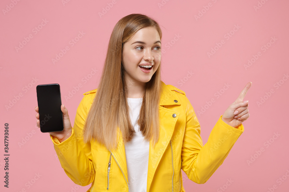 Fototapeta premium Funny young woman girl in yellow leather jacket isolated on pastel pink background. People lifestyle concept. Mock up copy space. Hold mobile phone with blank empty screen point index finger aside up.