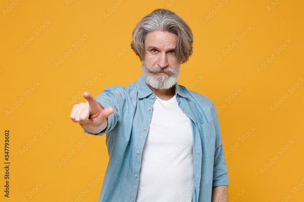 Strict elderly gray-haired mustache bearded man in casual blue shirt posing isolated on yellow wall background in studio. People lifestyle concept. Mock up copy space. Pointing index finger on camera. Strict elderly gray-haired mustache bearded man in casual blue shirt posing isolated on yellow wall background in studio. People lifestyle concept. Mock up copy space. Pointing index finger on camera.
