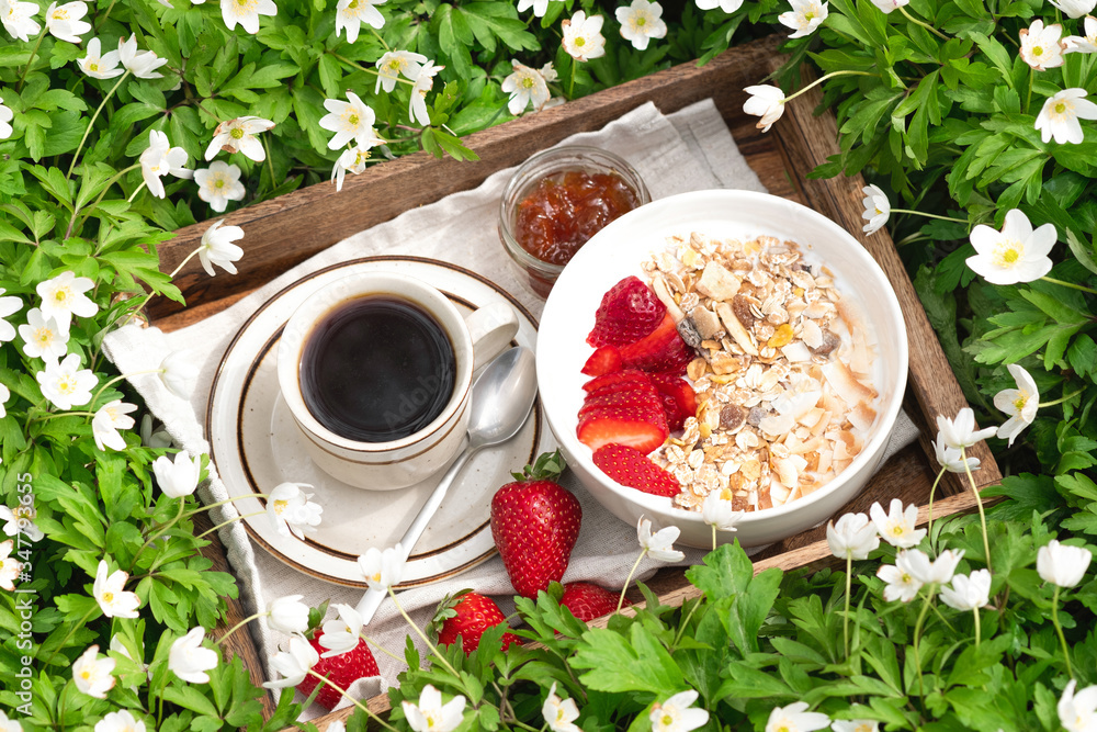 Summer picnic on a sunny day. Breakfast on green grass in the garden with white spring flowers blossom. Eating food outdoor. Cup of black coffee with bowl of yogurt and muesli with berries. Close up