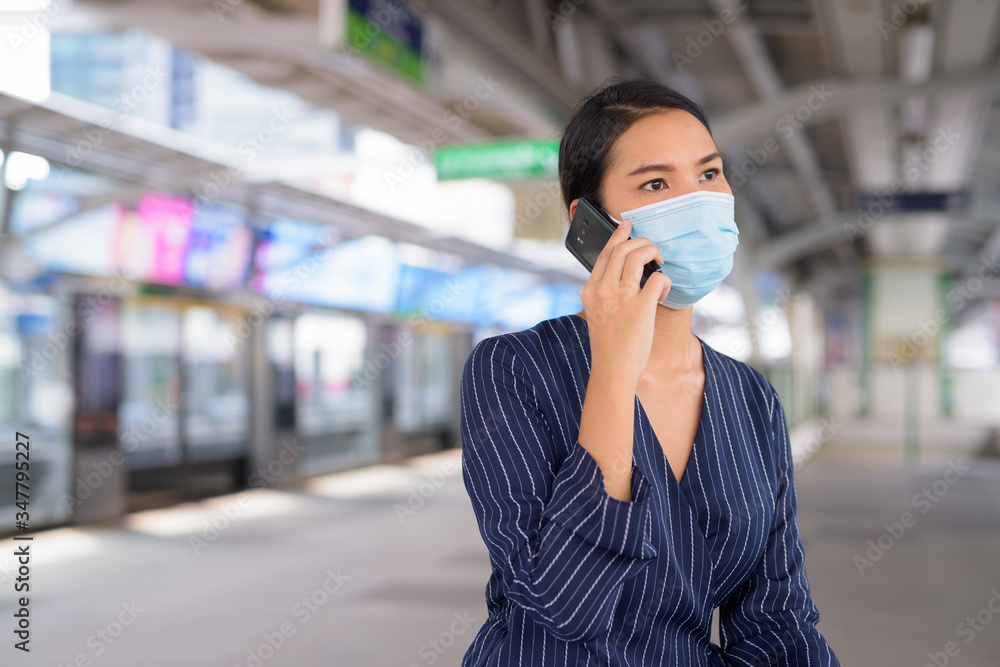 Young Asian businesswoman with mask talking on the phone while sitting at the skytrain station Young Asian businesswoman with mask talking on the phone while sitting at the skytrain station