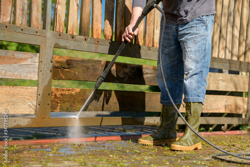 Unrecognizable man cleaning a wooden gate with a power washer. High water pressure cleaner used to DIY repair garden gate.