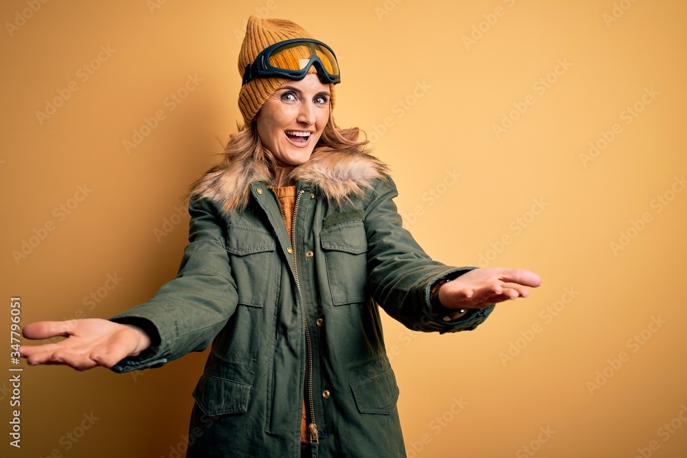 Middle age beautiful blonde skier woman wearing snow sportwear and ski goggles smiling cheerful offering hands giving assistance and acceptance.