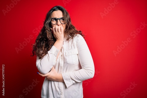Young beautiful woman with curly hair wearing shirt and glasses over red background looking stressed and nervous with hands on mouth biting nails. Anxiety problem.