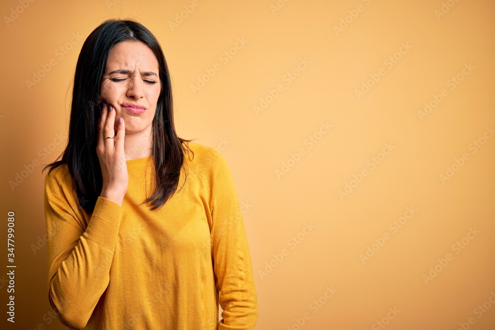 Young brunette woman with blue eyes wearing casual sweater over yellow background touching mouth with hand with painful expression because of toothache or dental illness on teeth. Dentist Young brunette woman with blue eyes wearing casual sweater over yellow background touching mouth with hand with painful expression because of toothache or dental illness on teeth. Dentist