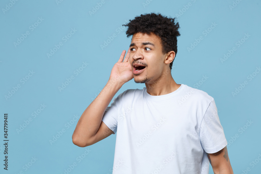 Curious young african american guy in casual white t-shirt posing isolated on pastel blue background studio portrait. People lifestyle concept. Mock up copy space. Try to hear you with hand near ear. Curious young african american guy in casual white t-shirt posing isolated on pastel blue background studio portrait. People lifestyle concept. Mock up copy space. Try to hear you with hand near ear.