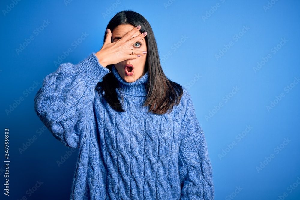 Young brunette woman with blue eyes wearing casual turtleneck sweater peeking in shock covering face and eyes with hand, looking through fingers with embarrassed expression. Young brunette woman with blue eyes wearing casual turtleneck sweater peeking in shock covering face and eyes with hand, looking through fingers with embarrassed expression.