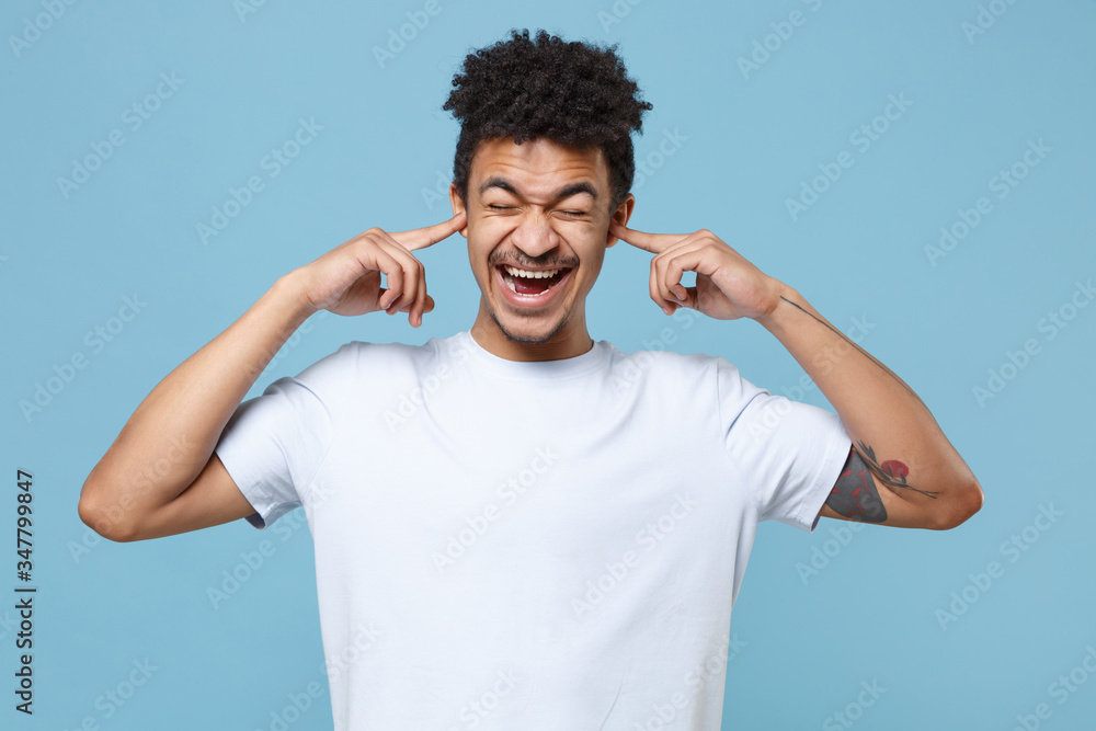Crazy young african american guy in casual white t-shirt posing isolated on blue background. People lifestyle concept. Mock up copy space. Covering ears with fingers, keeping eyes closed, screaming. Crazy young african american guy in casual white t-shirt posing isolated on blue background. People lifestyle concept. Mock up copy space. Covering ears with fingers, keeping eyes closed, screaming.