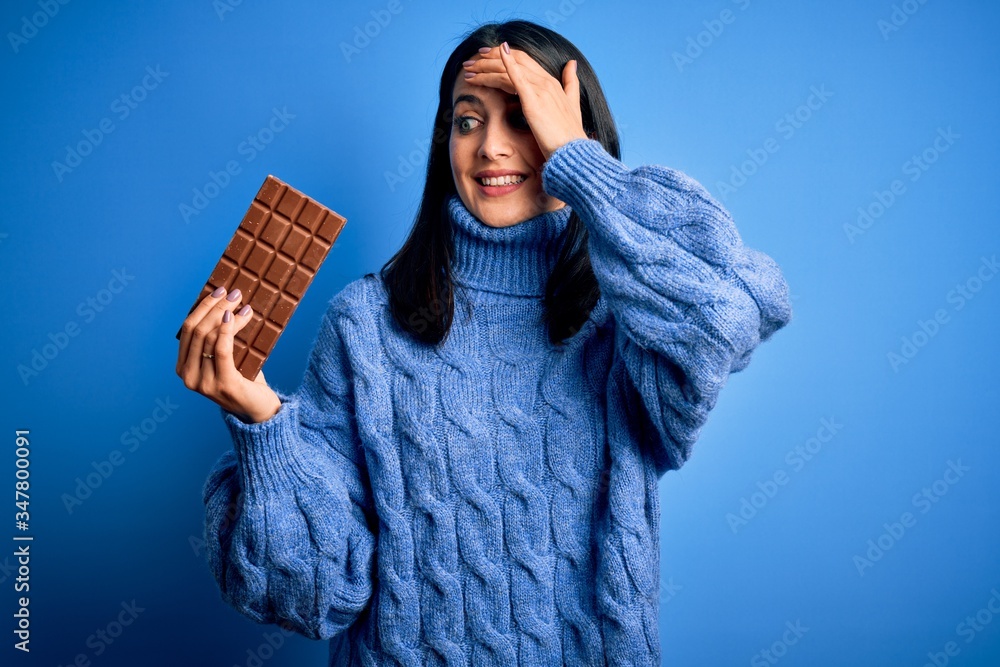 Young woman with blue eyes holding sweet chocolate bar standing over isolated background stressed with hand on head, shocked with shame and surprise face, angry and frustrated. Fear and upset Young woman with blue eyes holding sweet chocolate bar standing over isolated background stressed with hand on head, shocked with shame and surprise face, angry and frustrated. Fear and upset