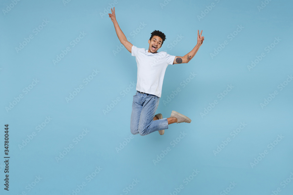 Cheerful young african american guy in casual white t-shirt posing isolated on pastel blue wall background studio portrait. People lifestyle concept. Mock up copy space. Jumping, showing victory sign.