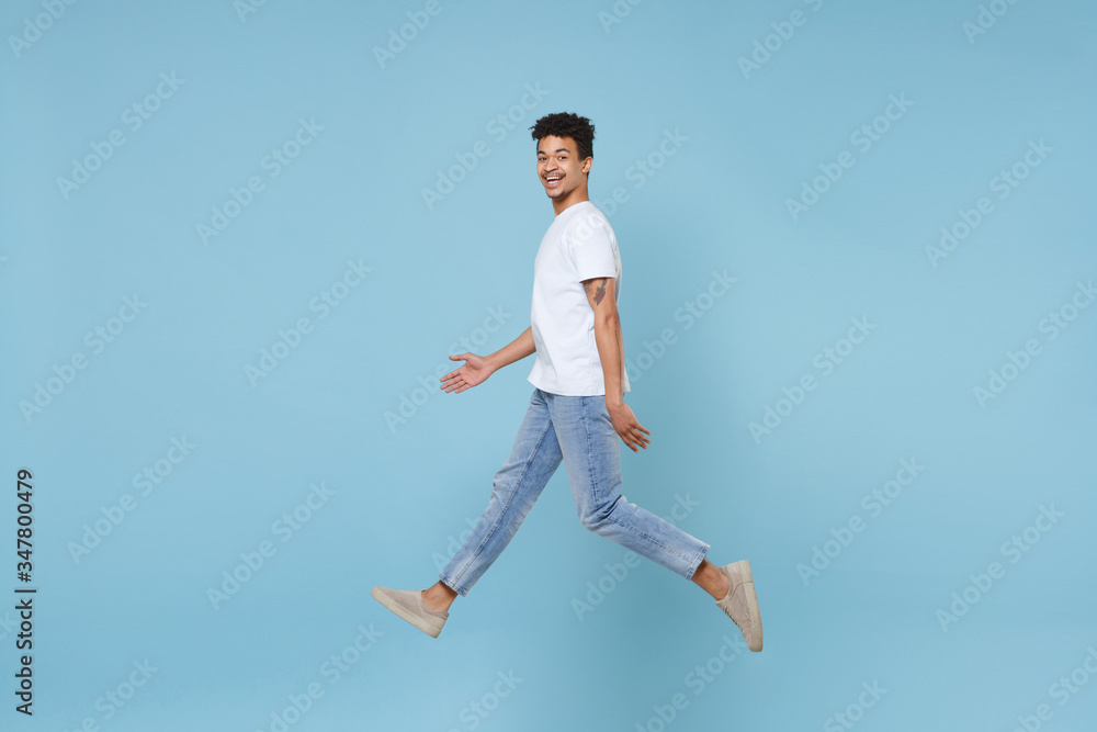 Side view of smiling young african american guy in casual white t-shirt posing isolated on blue background studio. People lifestyle concept. Mock up copy space. Jumping spreading hands looking camera. Side view of smiling young african american guy in casual white t-shirt posing isolated on blue background studio. People lifestyle concept. Mock up copy space. Jumping spreading hands looking camera.