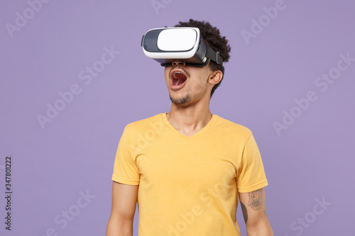 Shocked young african american guy in casual yellow t-shirt posing isolated on pastel violet background studio. People lifestyle concept. Mock up copy space. Watching in headset, keeping mouth open.