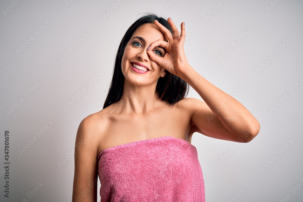 Fototapeta premium Young brunette woman with blue eyes wearing shower towel after bath over white background doing ok gesture with hand smiling, eye looking through fingers with happy face.
