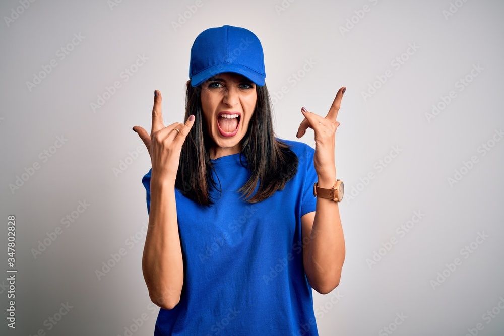 Young delivery woman with blue eyes wearing cap standing over blue background shouting with crazy expression doing rock symbol with hands up. Music star. Heavy concept. Young delivery woman with blue eyes wearing cap standing over blue background shouting with crazy expression doing rock symbol with hands up. Music star. Heavy concept.