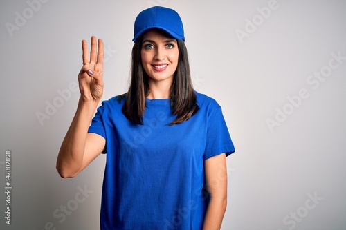 Young delivery woman with blue eyes wearing cap standing over blue background showing and pointing up with fingers number three while smiling confident and happy.