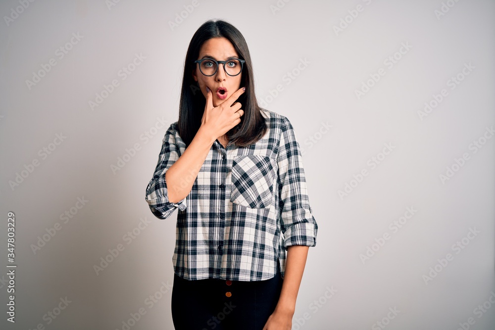 Young brunette woman with blue eyes wearing casual shirt and glasses over white background Looking fascinated with disbelief, surprise and amazed expression with hands on chin