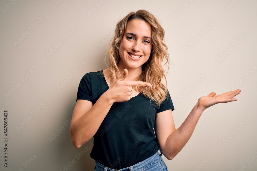 Young beautiful blonde woman wearing casual t-shirt standing over isolated white background amazed and smiling to the camera while presenting with hand and pointing with finger. Young beautiful blonde woman wearing casual t-shirt standing over isolated white background amazed and smiling to the camera while presenting with hand and pointing with finger.