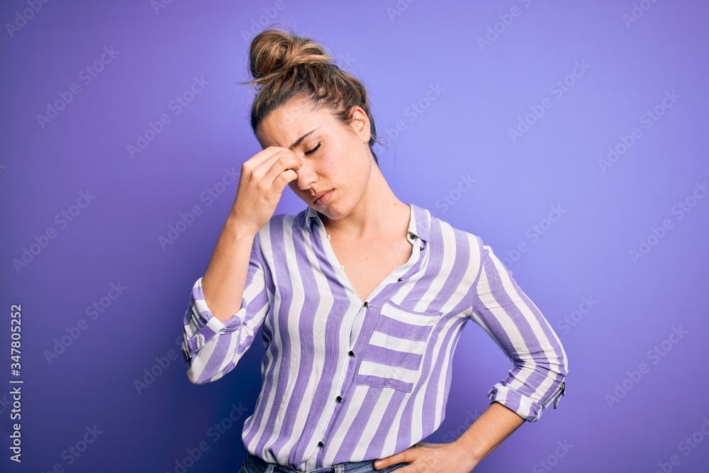 Young beautiful blonde woman wearing casual striped shirt standing over purple background tired rubbing nose and eyes feeling fatigue and headache. Stress and frustration concept.