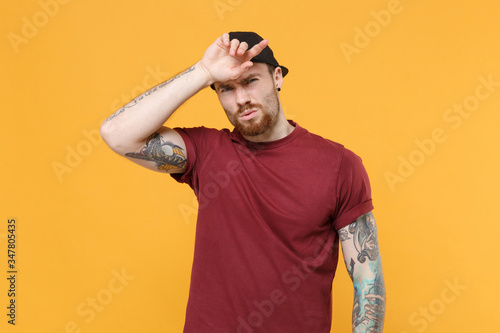 Exhausted tired young bearded tattooed man guy in casual t-shirt, black cap posing isolated on yellow wall background studio portrait. People lifestyle concept. Mock up copy space. Put hands on head. photo