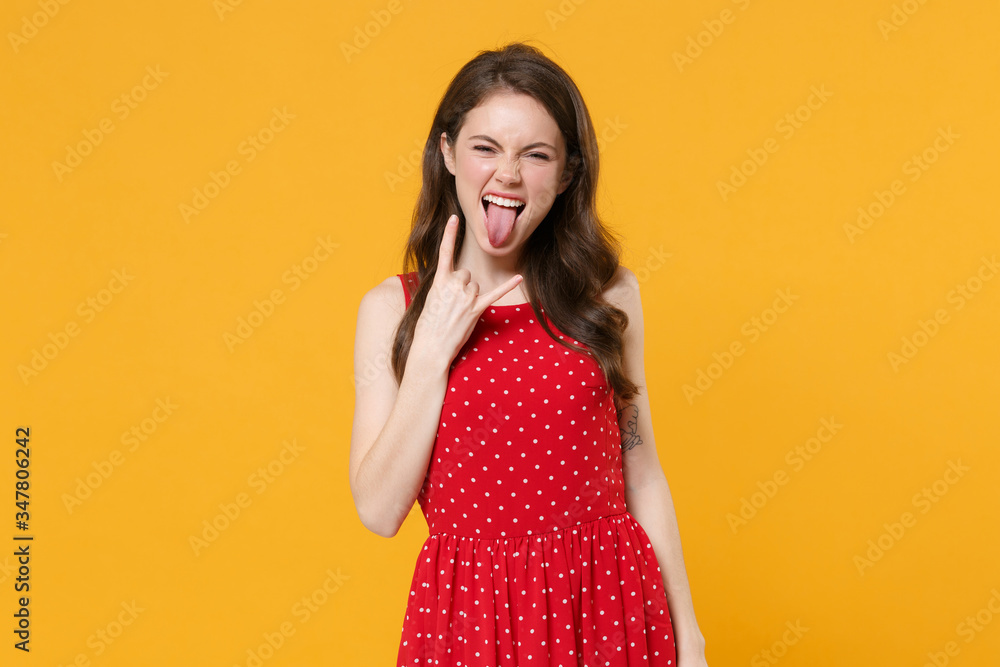 Funny young brunette woman girl in red summer dress posing isolated on yellow background. People lifestyle concept. Mock up copy space. Depicting heavy metal rock sign horns up gesture showing tongue. Funny young brunette woman girl in red summer dress posing isolated on yellow background. People lifestyle concept. Mock up copy space. Depicting heavy metal rock sign horns up gesture showing tongue.