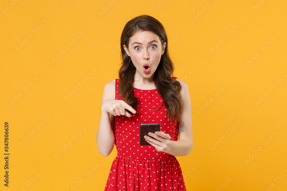 Shocked young brunette woman girl in red summer dress posing isolated on yellow wall background studio portrait. People lifestyle concept. Mock up copy space. Pointing index finger on mobile phone. Shocked young brunette woman girl in red summer dress posing isolated on yellow wall background studio portrait. People lifestyle concept. Mock up copy space. Pointing index finger on mobile phone.
