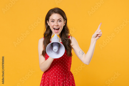 Excited young brunette woman girl in red summer dress posing isolated on yellow background in studio. People lifestyle concept. Mock up copy space. Scream in megaphone, pointing index finger aside up.