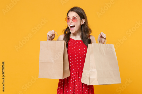 Surprised young brunette woman girl in red summer dress, eyeglasses posing isolated on yellow wall background studio portrait. People lifestyle concept. Hold package bag with purchases after shopping.