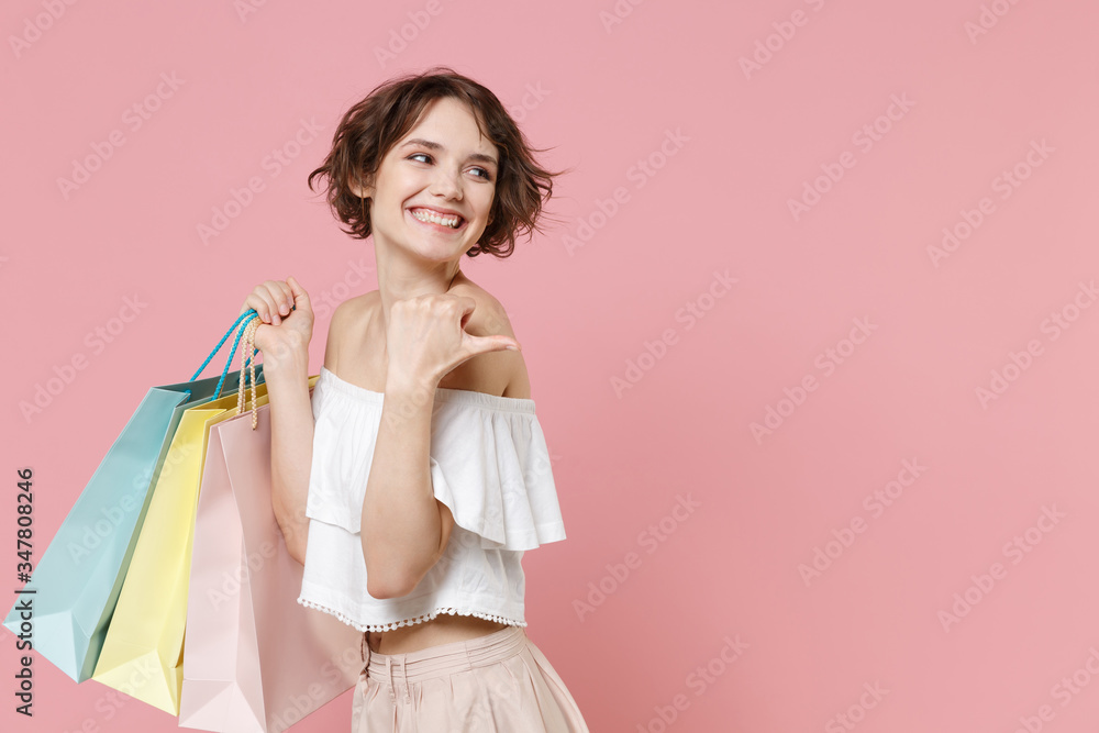 Side view of smiling young woman girl in summer clothes hold package bag with purchases isolated on pink background studio. Shopping discount sale concept. Mock up copy space. Pointing thumb aside.
