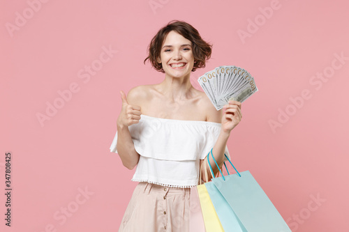 Smiling young woman girl in summer clothes isolated on pink wall background. Shopping discount sale concept. Hold package bag with purchases, fan of cash money in dollar banknotes, showing thumb up. © ViDi Studio