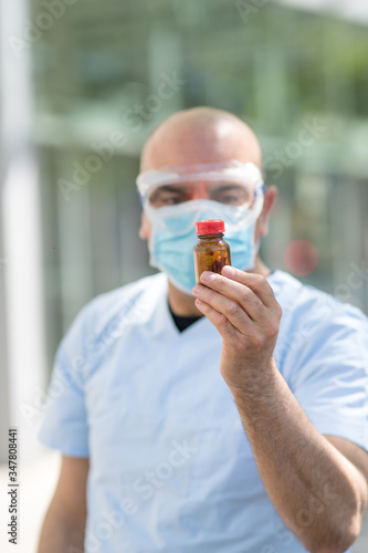 Portrait of an out of focused doctor wearing white coat, surgical mask and medical safety goggles holding a bottle of pills in his hand. Selective focus