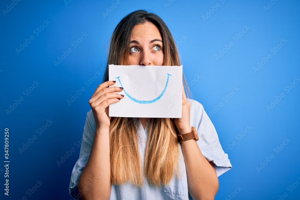 Young beautiful brunette woman holding card with smile illustration on mouth serious face thinking about question, very confused idea Young beautiful brunette woman holding card with smile illustration on mouth serious face thinking about question, very confused idea