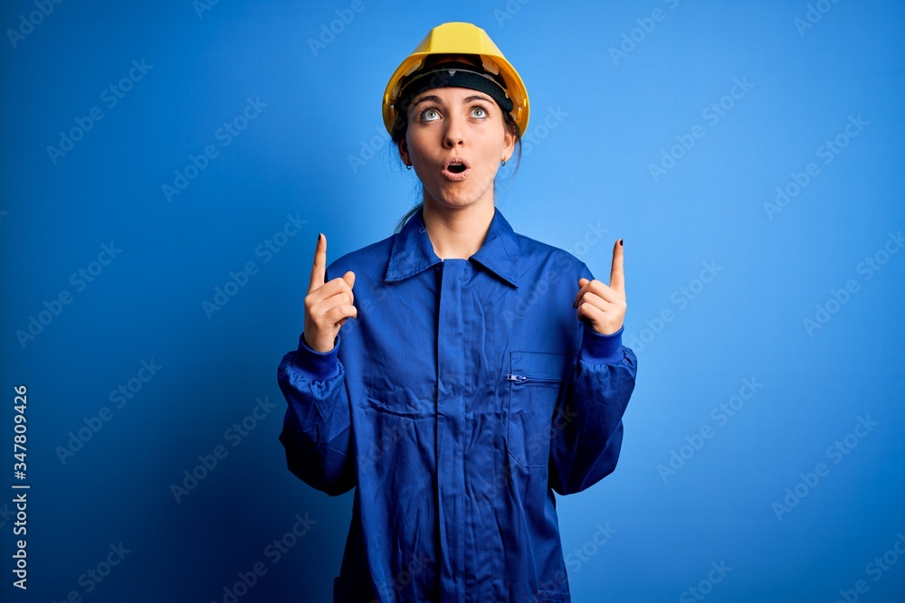 Young beautiful worker woman with blue eyes wearing security helmet and uniform amazed and surprised looking up and pointing with fingers and raised arms.