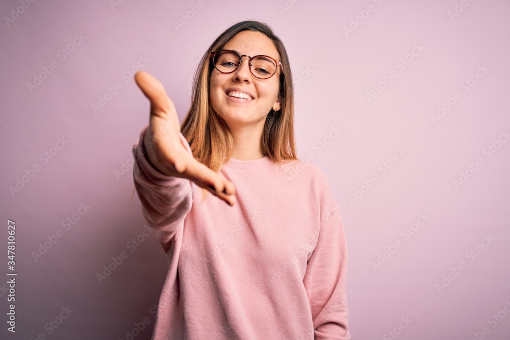 Beautiful blonde woman with blue eyes wearing sweater and glasses over pink background smiling friendly offering handshake as greeting and welcoming. Successful business. Beautiful blonde woman with blue eyes wearing sweater and glasses over pink background smiling friendly offering handshake as greeting and welcoming. Successful business.
