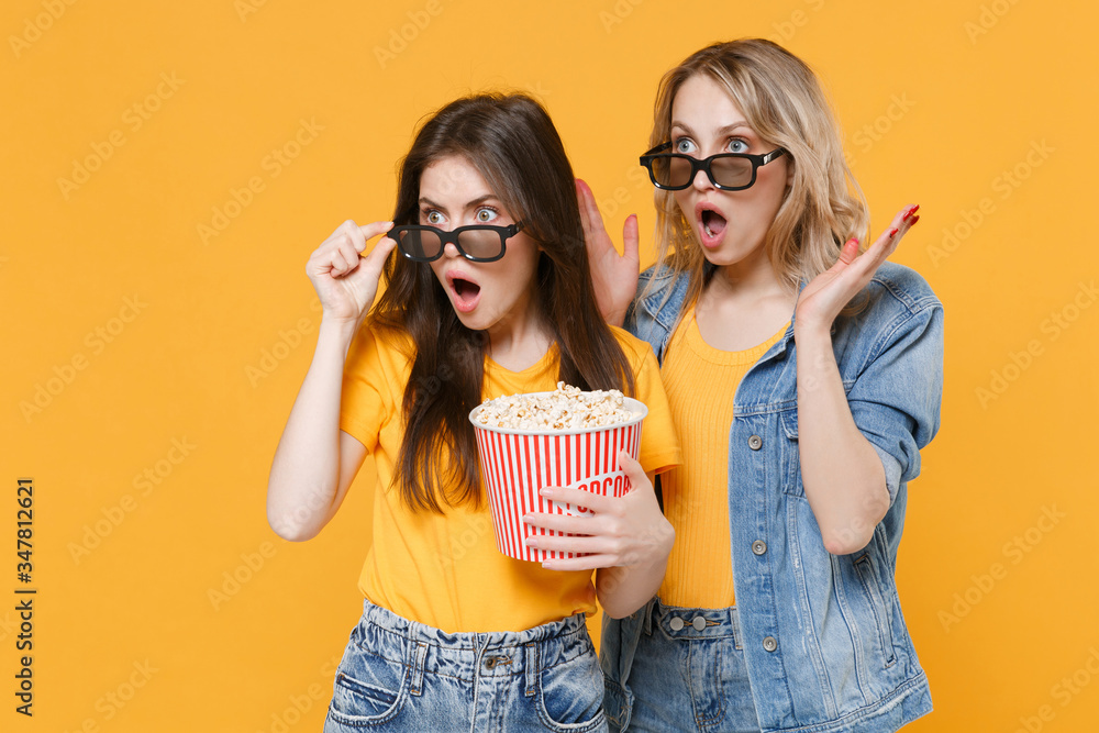Obraz premium Shocked concerned young women girls friends in denim clothes, 3d glasses isolated on yellow background. People sincere emotions in cinema lifestyle concept. Watching movie film hold bucket of popcorn.