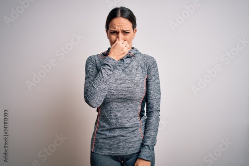 Young blonde fitness woman wearing sport workout clothes over isolated background smelling something stinky and disgusting, intolerable smell, holding breath with fingers on nose. Bad smell
