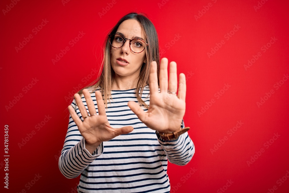 Young beautiful blonde woman with blue eyes wearing glasses standing over red background Moving away hands palms showing refusal and denial with afraid and disgusting expression. Stop and forbidden. Young beautiful blonde woman with blue eyes wearing glasses standing over red background Moving away hands palms showing refusal and denial with afraid and disgusting expression. Stop and forbidden.