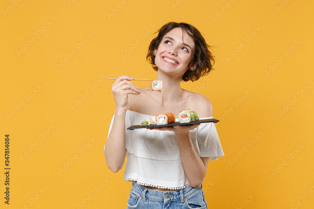 Young brunette woman girl in casual clothes hold in hand makizushi sushi roll served on black plate traditional japanese food isolated on yellow background studio portrait. People lifestyle concept.