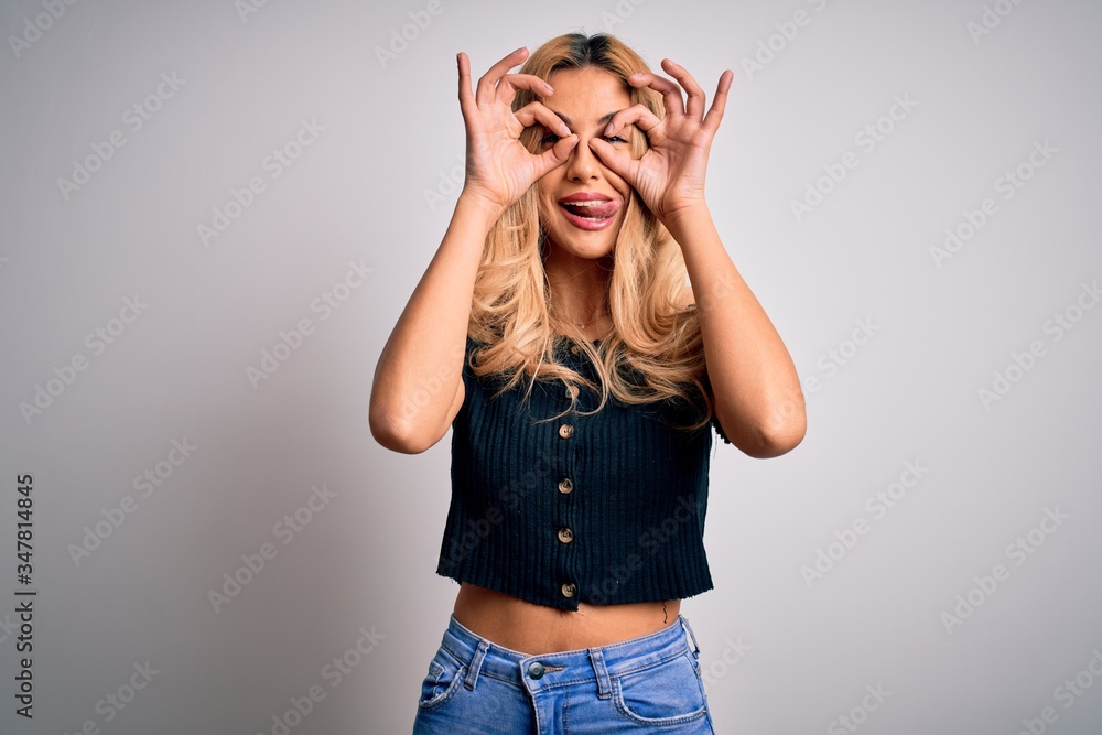 Young beautiful blonde woman wearing casual t-shirt standing over isolated white background doing ok gesture like binoculars sticking tongue out, eyes looking through fingers. Crazy expression. Young beautiful blonde woman wearing casual t-shirt standing over isolated white background doing ok gesture like binoculars sticking tongue out, eyes looking through fingers. Crazy expression.