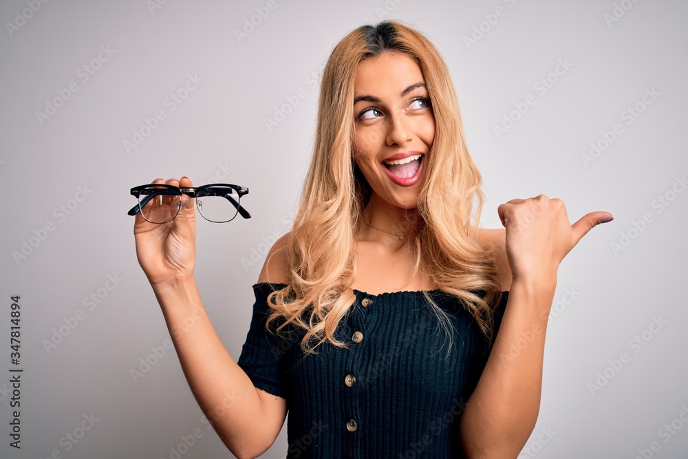 Young beautiful blonde optical woman holding vision glasses over isolated white background pointing and showing with thumb up to the side with happy face smiling Young beautiful blonde optical woman holding vision glasses over isolated white background pointing and showing with thumb up to the side with happy face smiling