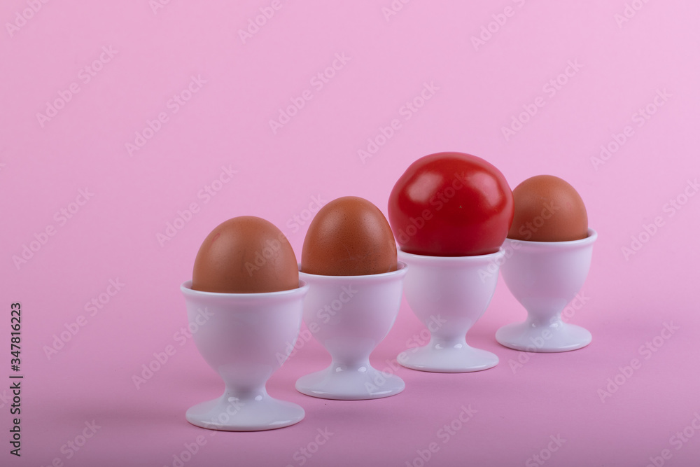 three brown eggs and a tomato in white eggs cups before pink background three brown eggs and a tomato in white eggs cups before pink background