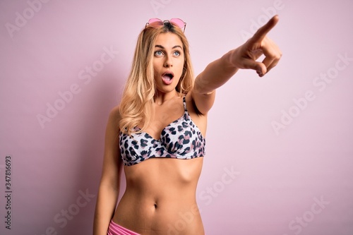 Young beautiful blonde woman on vacation wearing bikini over isolated pink background Pointing with finger surprised ahead, open mouth amazed expression, something on the front