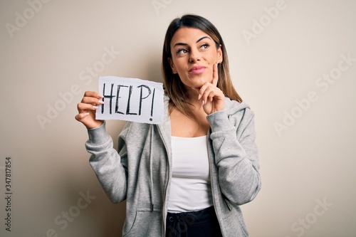 Young beautiful brunette woman holding paper with help message over white background serious face thinking about question, very confused idea
