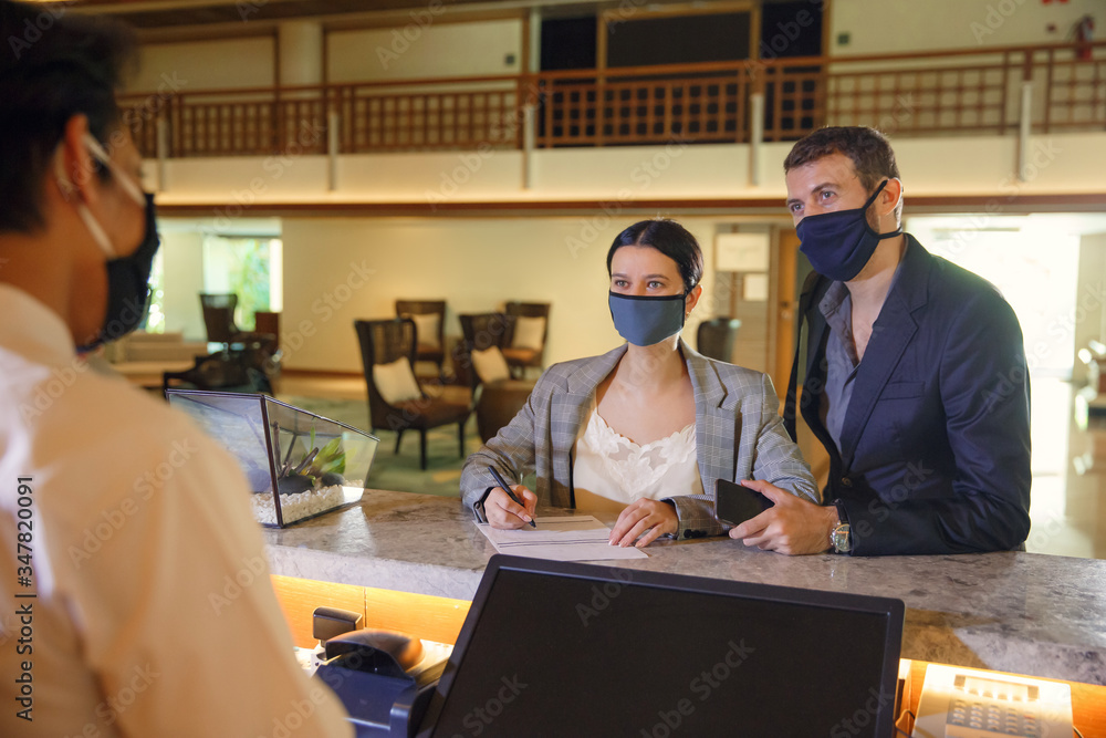Fototapeta premium Couple and receptionist at counter in hotel wearing medical masks as precaution against virus. Couple on a business trip doing check-in at the hotel