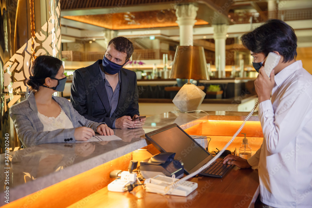 Couple and receptionist at counter in hotel wearing medical masks as precaution against virus. Couple on a business trip doing check-in at the hotel