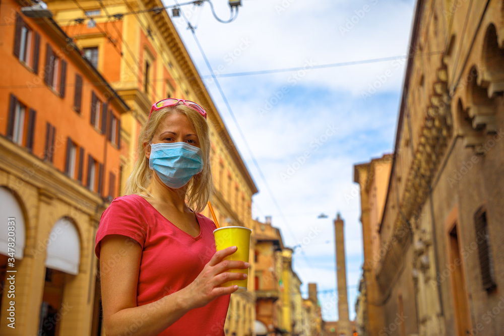 COVID-19 Italian quarantine: blonde girl with surgical mask having a drink on the road with Two Towers of Bologna. Coronavirus after quarantine with social distancing in Bologna city of Italy. COVID-19 Italian quarantine: blonde girl with surgical mask having a drink on the road with Two Towers of Bologna. Coronavirus after quarantine with social distancing in Bologna city of Italy.