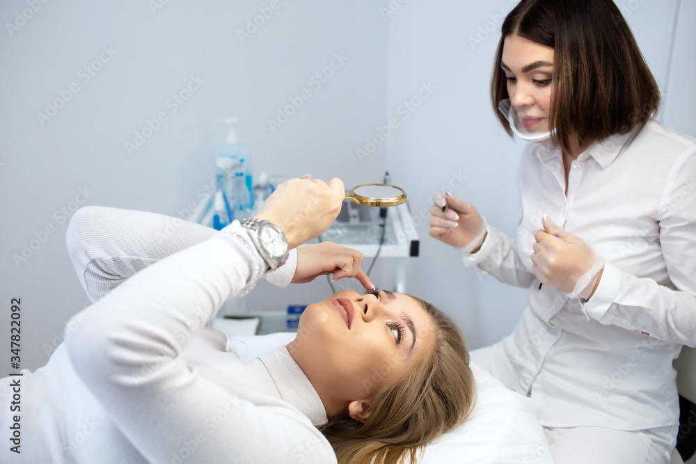 Beautiful girl patient looks in the mirror during a facial skin care procedure. The use of permanent makeup on brows. The wizard works with eyebrows, shows the results of work. Semi-permanent makeup. Beautiful girl patient looks in the mirror during a facial skin care procedure. The use of permanent makeup on brows. The wizard works with eyebrows, shows the results of work. Semi-permanent makeup.