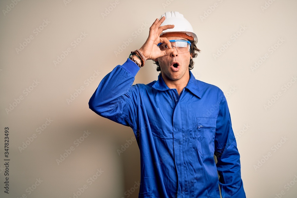 Young constructor man wearing uniform and security helmet over isolated white background doing ok gesture shocked with surprised face, eye looking through fingers. Unbelieving expression.