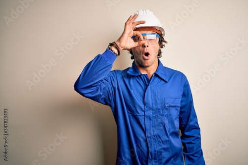 Young constructor man wearing uniform and security helmet over isolated white background doing ok gesture shocked with surprised face  eye looking through fingers. Unbelieving expression.